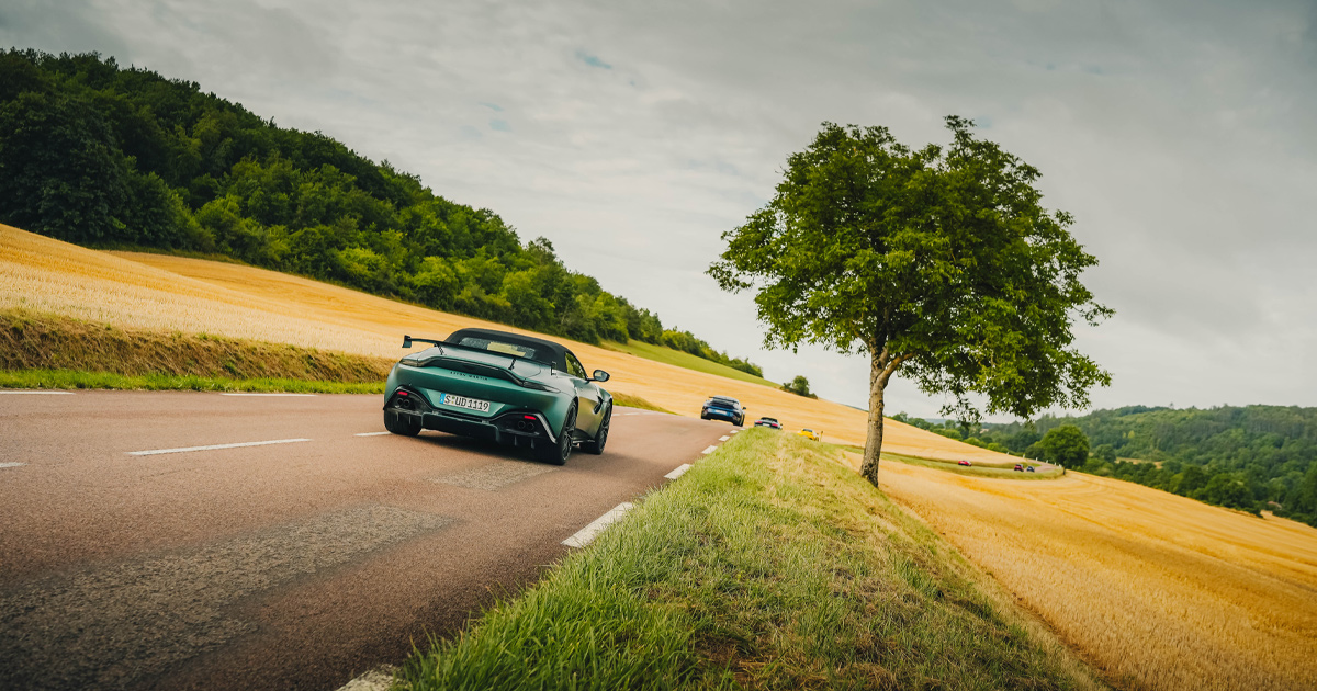 A green Aston Martin Vantage convertible with black hood driving through golden French farmlands