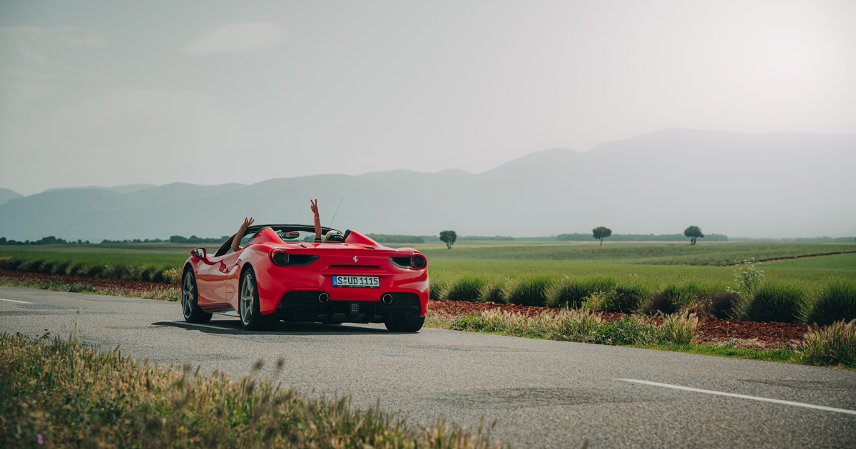 A red Ferrari 488 Spyder with driver and passenger’s hands raised aloft on a country road in France