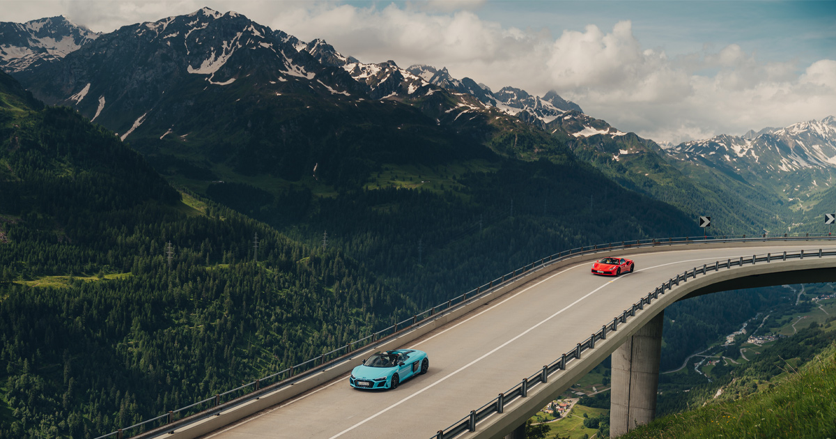 A blue Audi R8 leads a red Lamborghini Huracan along a high alpine road in Europe