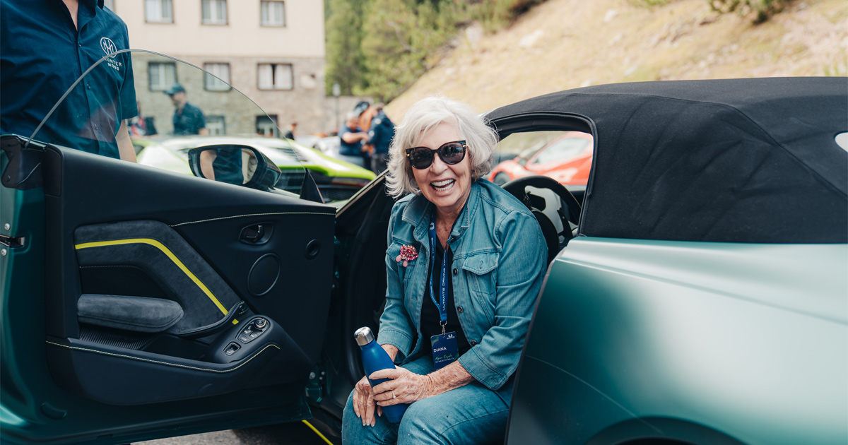 A woman in sunglasses smiles as she sits in the driver’s seat of an Aston Martin