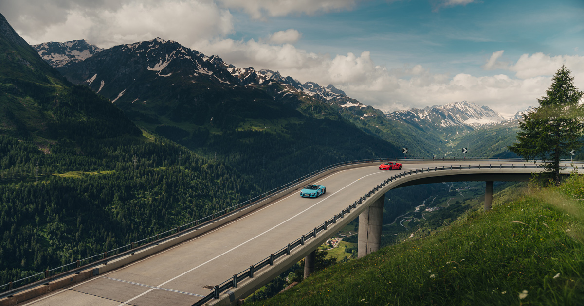 A blue Audi R8 leads a red Lamborghini along a bridge in the Alps 