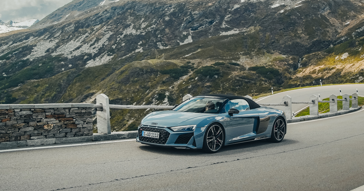 A blue Audi R8 Spyder with roof up on a high alpine road exiting a tight corner