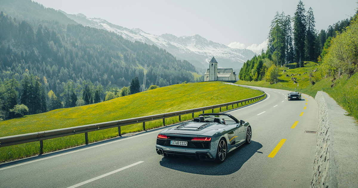 A blue Audi R8 Spyder behind a Porsche 911 on a mountain valley road in Europe 