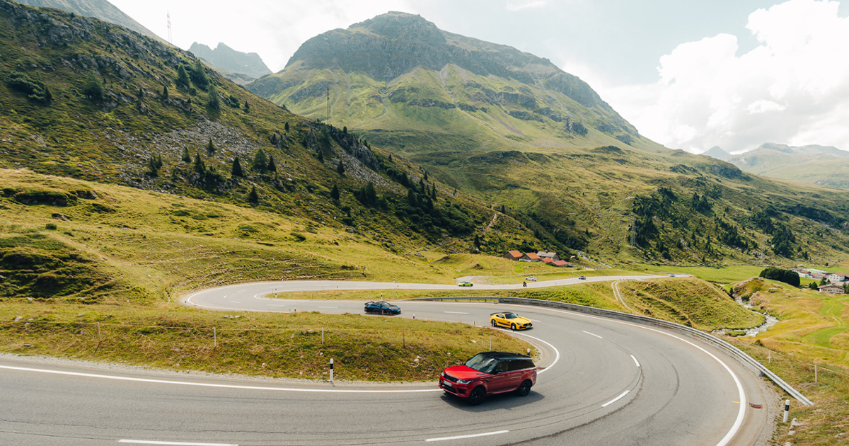 A red Range Rover leads a supercar fleet along a twisty road in the hills