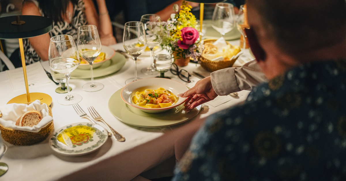 A man is served a bowl of filled pasta in broth at a table with white table cloth and wine glasses