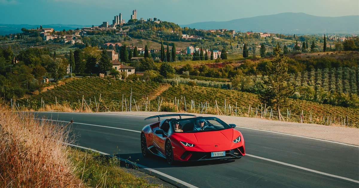 A red Lamborghini Huracan Spyder rounds a corner in the Tuscan hills surrounded by vineyards.