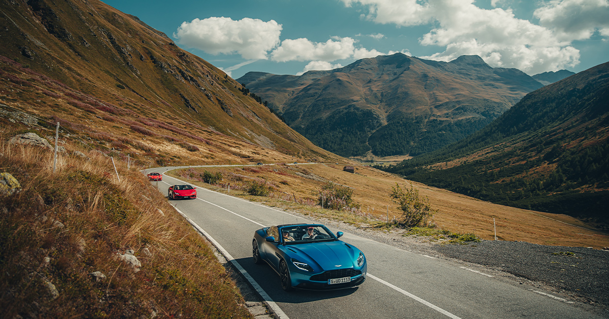 A green Aston Martin Vantage convertible leads two red supercars along an epic country road